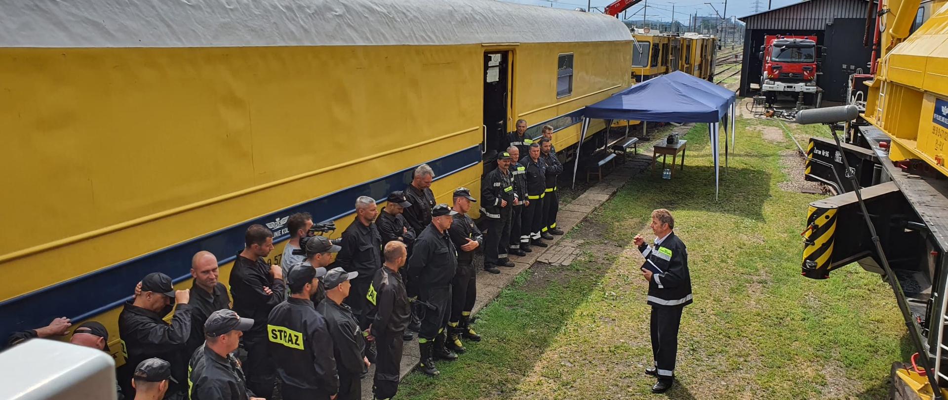 Strażacy w ubraniu koszarowym stoją na tle żółtego wagonu pociągu. Na przeciw pracownik ratownictwa kolejowego.