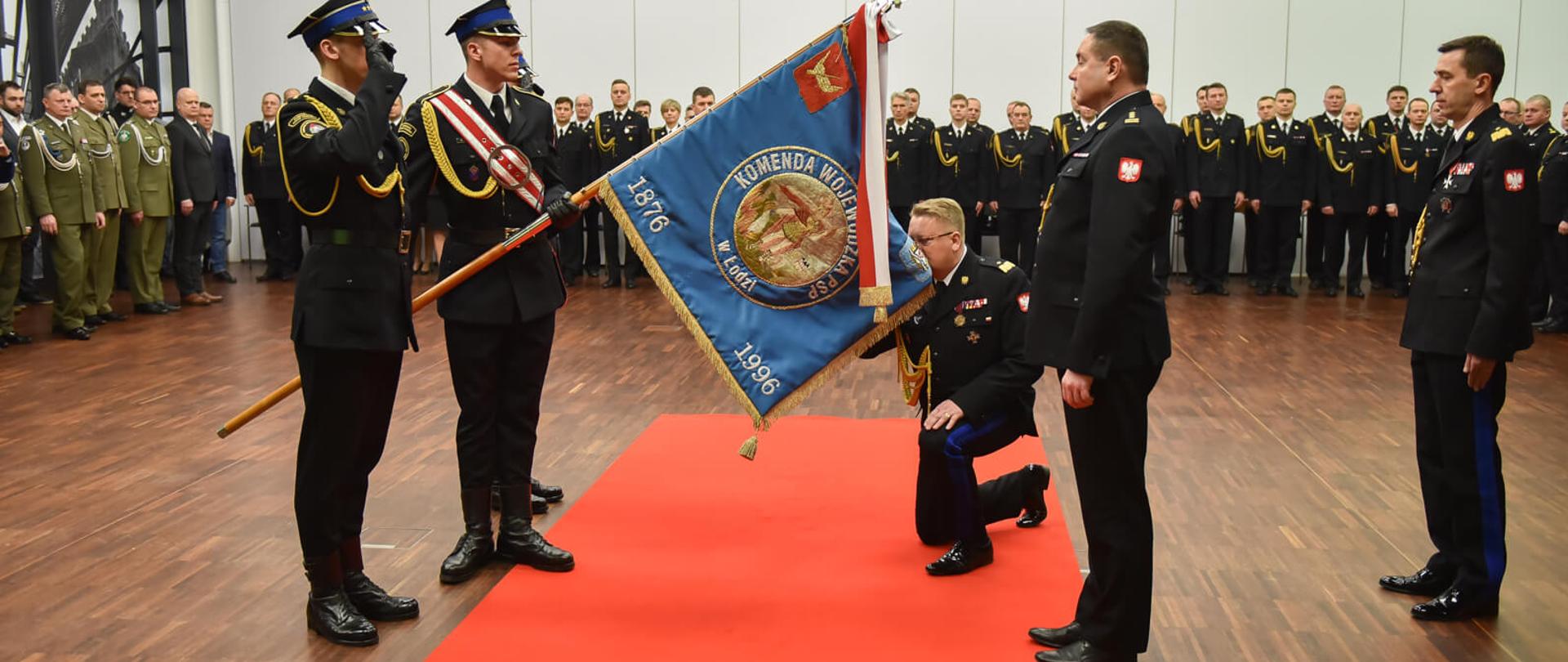 nadbrygadier Grzegorz Janowski przyklęka i całuje sztandar komendy wojewódzkiej po lewej stoi poczet sztandarowy po prawej dwóch strażaków, w tle uczestnicy uroczystości