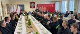 Przy stołach na których znajdują się dekoracje świąteczne i talerze, siedzą strażacy i zaproszenie goście. Strażacy ubrani są w mundury galowe zaproszeni goście odświętnie w garniturach. W tle logo Państwowej Straży Pożarnej oraz godło Polski i flagi
