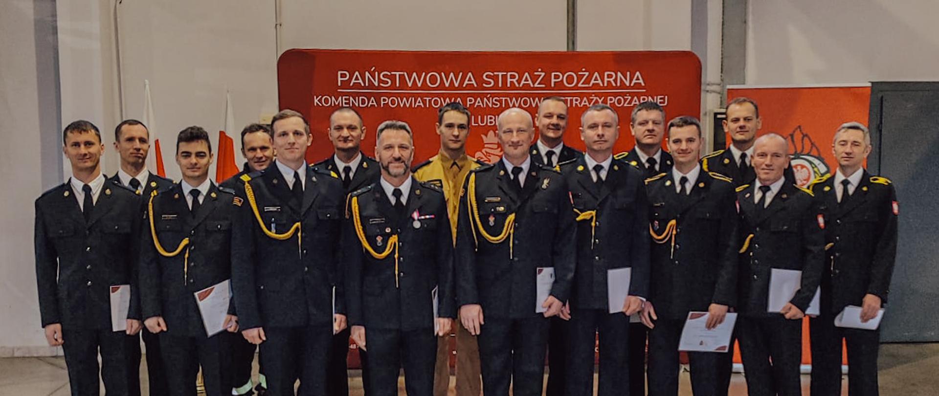 Zdjęcie zrobione w garażu strażnicy. Na pierwszym planie grupa strażaków w ubraniach galowych, skierowani twarzami w stronę obiektywu. W tle ścianka koloru czerwonego z widocznym napisem „Państwowa Straż Pożarna”