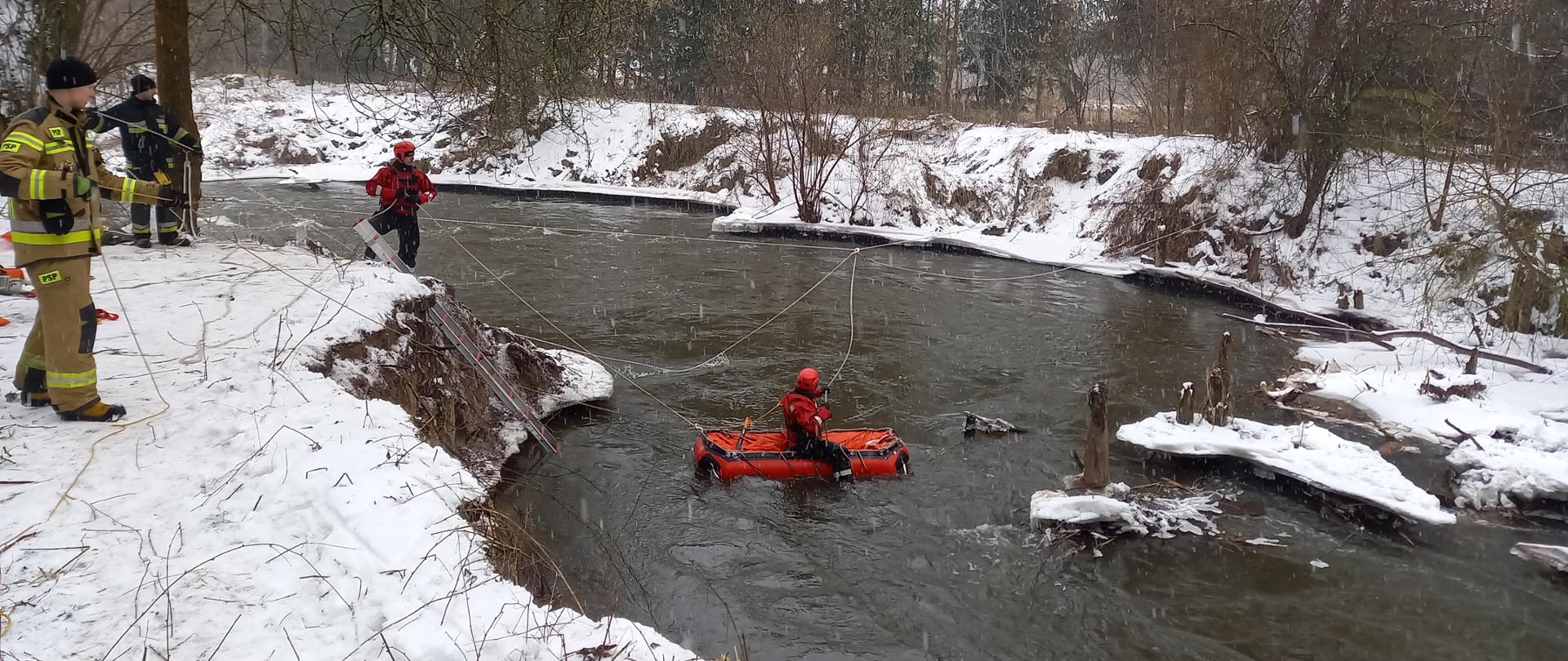 Na zdjęciu widoczni strażacy ubrani w czerwone stroje do ratownictwa wodnego. Jeden ratownik płynie na pontonie po rzece, drugi asekuruje go z brzegu. Z lewej strony widoczni strażacy ubrani w nomexy asekurujący osobę w wodzie. 