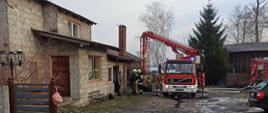 Zdjęcie w ciągu dnia. Na zdjęciu widać pojazd ratowniczo-gaśniczy w kolorze czerwonym, posiada sygnalizację ostrzegawczą. Zdjęcie w pobliżu budynku, w którym doszło do pożaru. Widać ratowników w ubraniu specjalnym.