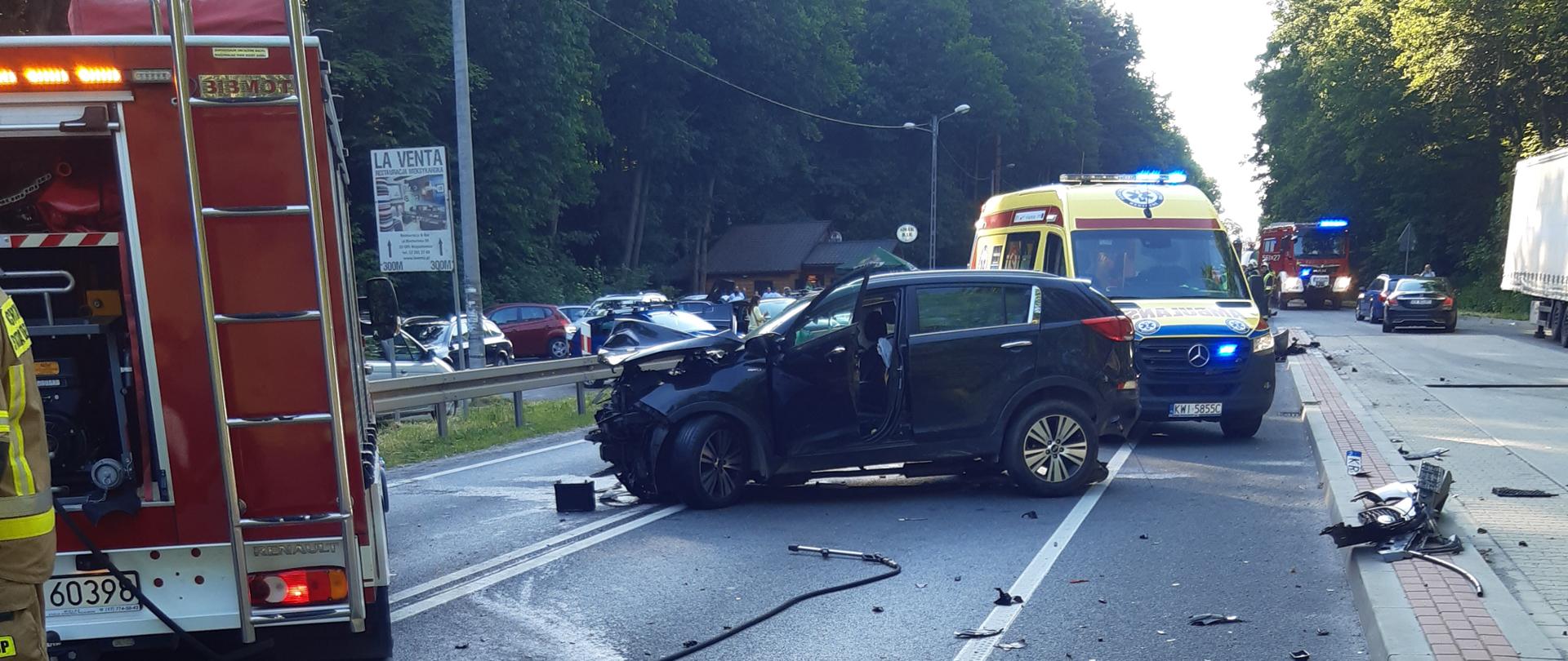 Na zdjęciu widoczny samochód stojący w poprzek drogi. Rozbity przód auta. Przez autem soi wóz strażacki. Za autem karetka.