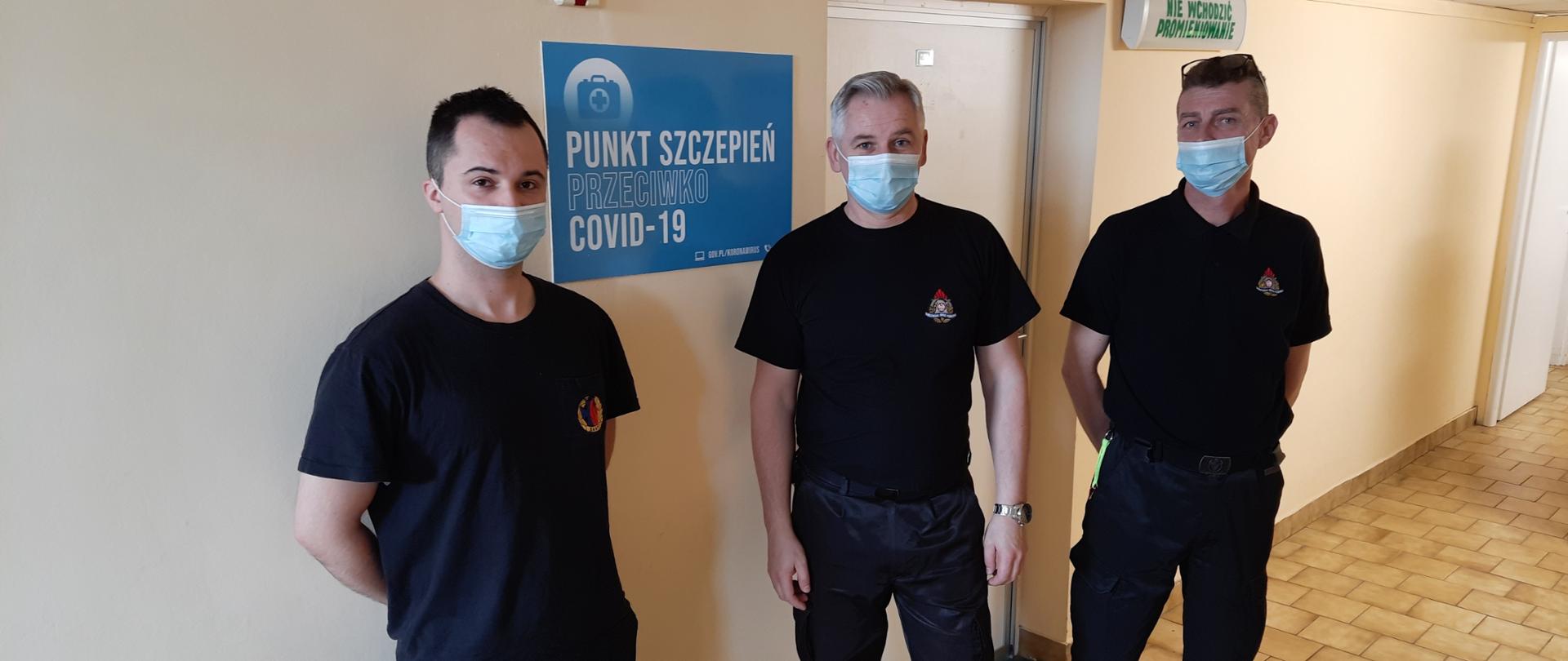 Zdjęcie przedstawia trzech funkcjonariuszy Komendy Powiatowej Państwowej Straży Pożarnej w Lwówku Śląskim stojących na korytarzu przed wejściem do punktu szczepień.