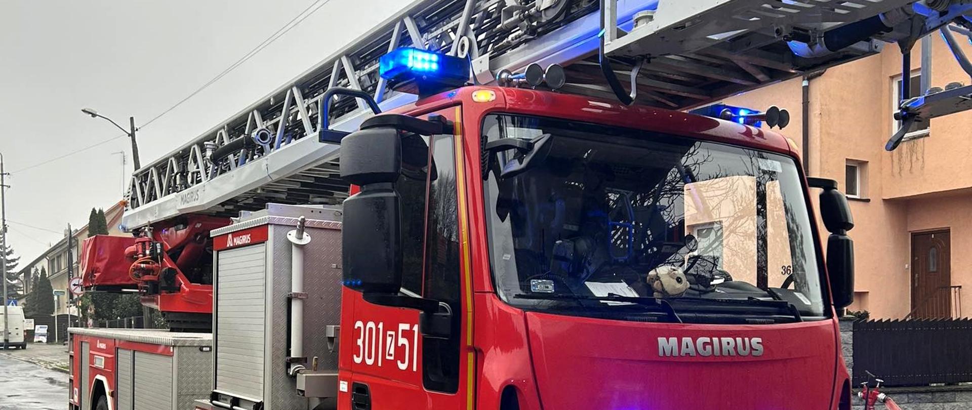 Na pierwszym planie samochód pożarniczy z drabiną mechaniczną na ulicy.