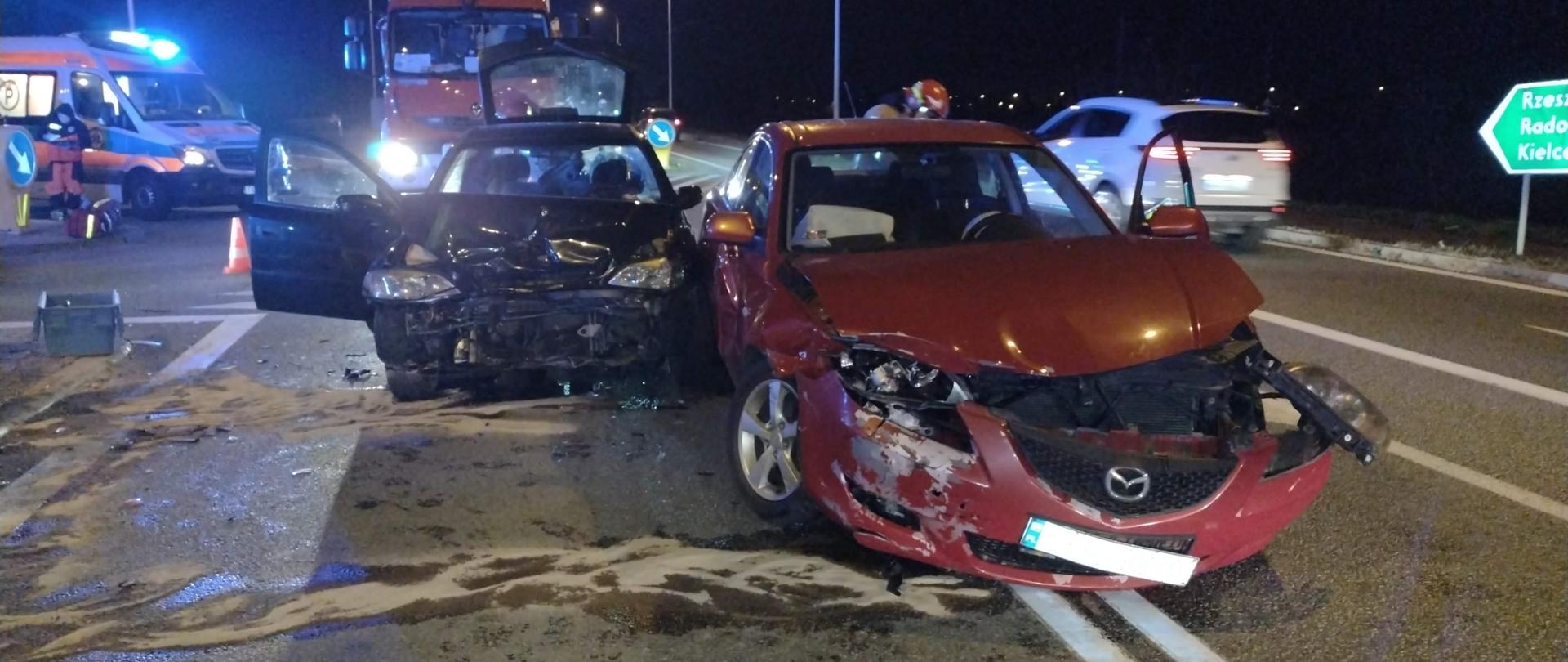 Na zdjęciu stoją przodem dwa rozbite auta osobowe. W tle pojazdy służb medycznych. Pora nocna.