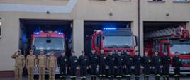 strażacy stoją w szeregu na baczność, za nimi wozy strażackie z włączonymi sygnałami świetlnymi
