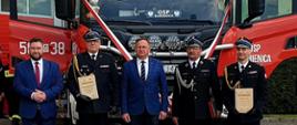 Na tle samochodu pożarniczego stoi w szeregu pięć osób, trzy z nich to strażacy w umundurowaniu galowym.