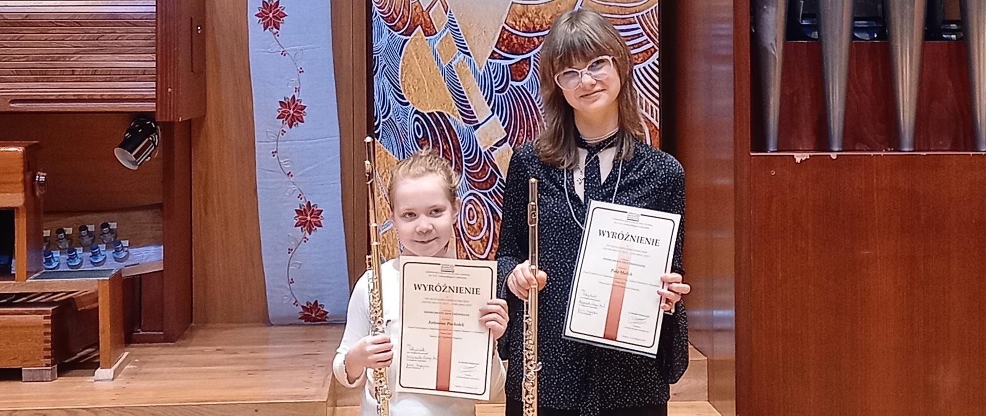 Uczennice ZPSM trzymają w ręku dyplomy i flety poprzeczne, stoją na tle organów i baneru szkoły.