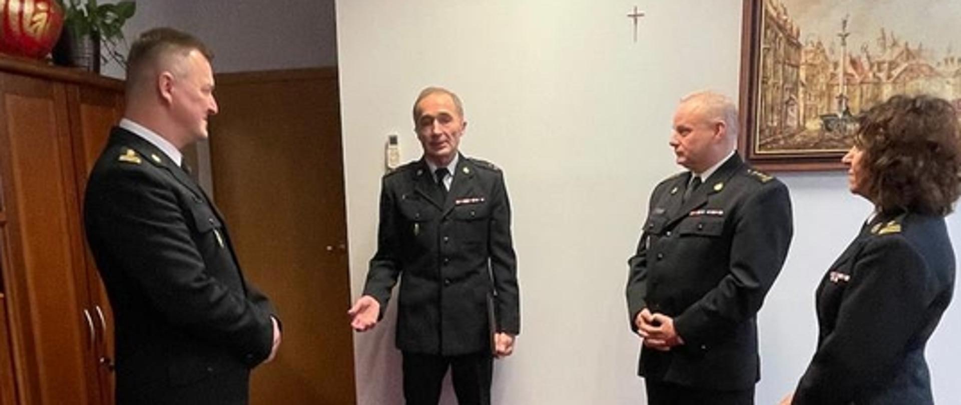 Na zdjęciu widać spotkanie czterech strażaków w mundurach służbowych w gabinecie komendanta. Jeden z nich coś opowiada, a pozostali go uważnie słuchają. Wśród zgromadzonych jest troje mężczyzn i jedna kobieta. Wszyscy stoją na czerwonej wykładzinie. W tle na ścianie obraz przedstawiający warszawską starówkę.