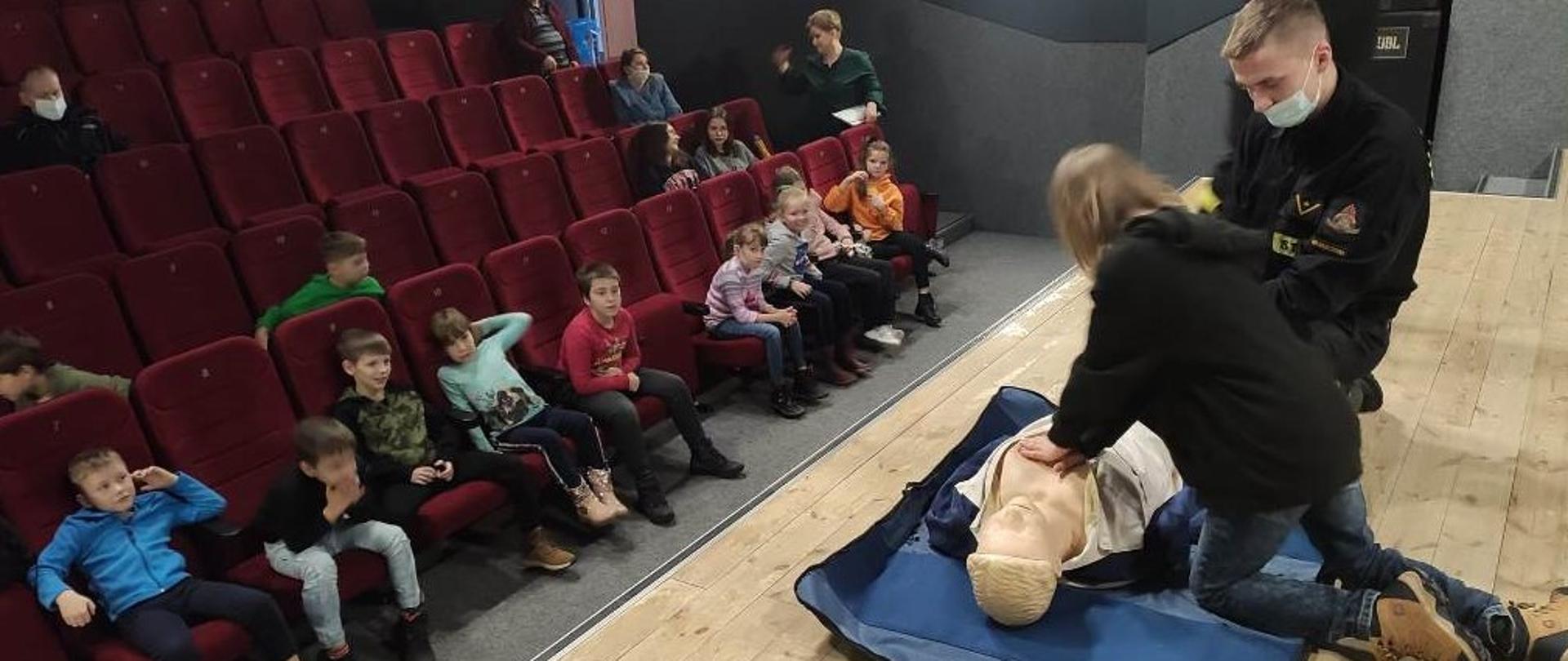 Dom Kultury, scena dużej Sali kinowej, obok strażaka jedna z uczestniczek spotkania uczy się prowadzić resuscytację krążeniowo-oddechową na fantomie. W lewej części zdjęcia uczestnicy spotkania siedzący na widowni.