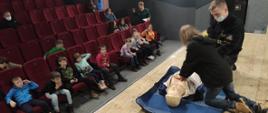 Dom Kultury, scena dużej Sali kinowej, obok strażaka jedna z uczestniczek spotkania uczy się prowadzić resuscytację krążeniowo-oddechową na fantomie. W lewej części zdjęcia uczestnicy spotkania siedzący na widowni.