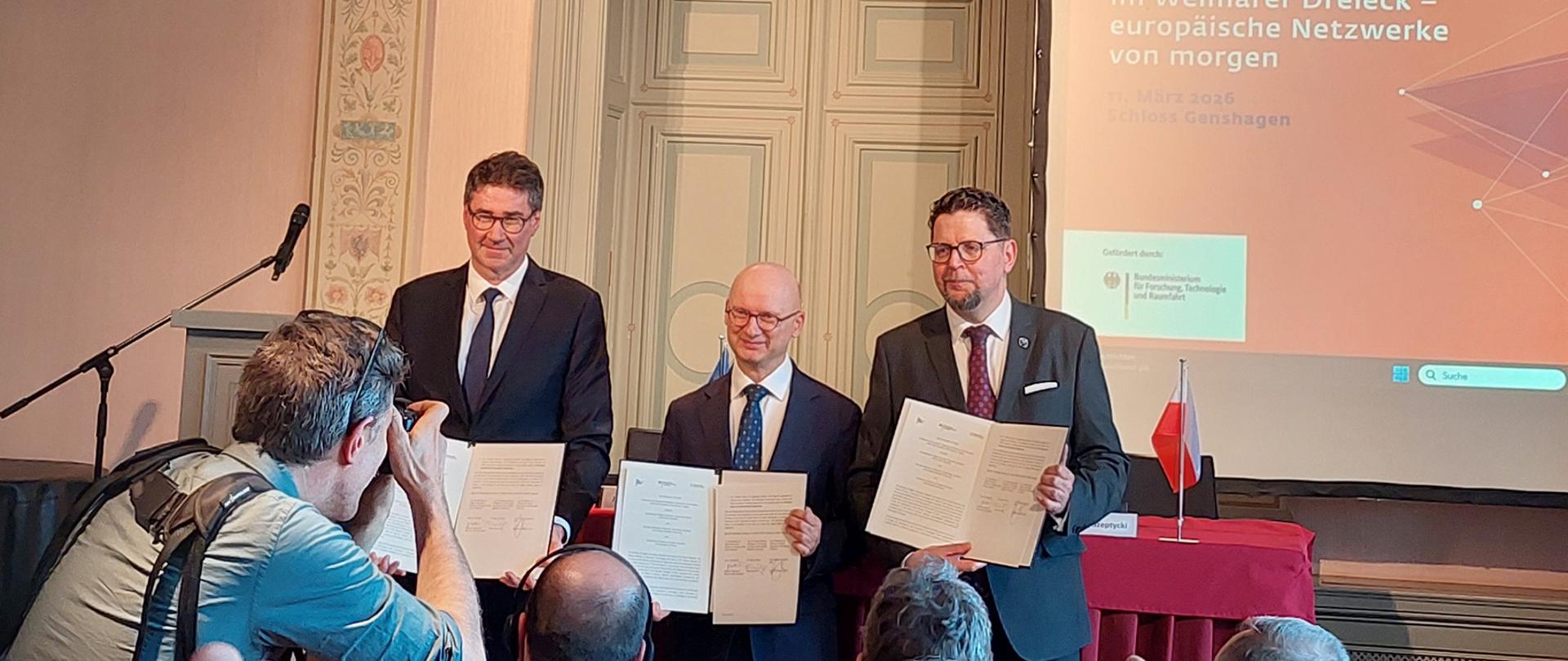 W wielkiej sali z różowymi ścianami przed nakrytym czerwonym materiałem stołem stoi trzech mężczyzn w garniturach, wiceminister Szeptycki pierwszy od prawej, pokazują trzymane w rękach otwarte teczki z dokumentami.