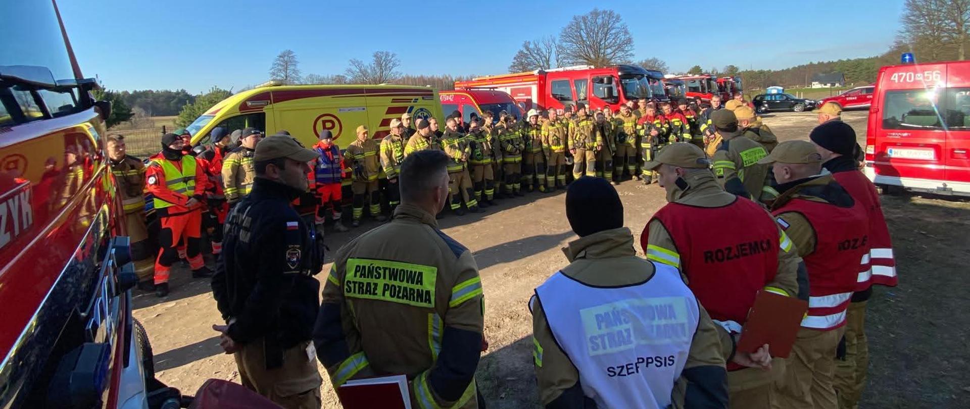 Na zdjęciu widać dużą grupę służb ratowniczych zgromadzonych na otwartym terenie, prawdopodobnie podczas odprawy lub koordynacji akcji. Obecni są strażacy oraz ratownicy medyczni w charakterystycznych mundurach i kamizelkach. W tle stoi wiele pojazdów służbowych – wozy strażackie, karetki oraz pojazdy operacyjne. Pogoda jest słoneczna, niebo bezchmurne, co sugeruje, że działania odbywają się w ciągu dnia. Całość sprawia wrażenie dużej, zorganizowanej akcji ratowniczej lub ćwiczeń służb.