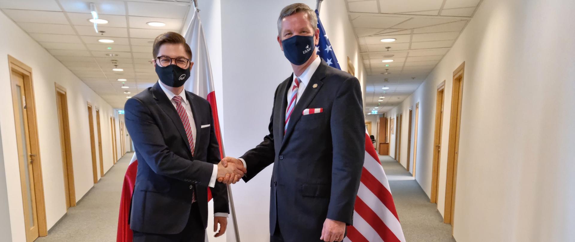 Spotkanie Prezesa Państwowej Agencji Atomistyki z Przewodniczącym Nuclear Regulatory Commission. Dr Łukasz Młynarkiewicz i Chistopher T. Hanson podają sobie ręce. W tle flaga polska i amerykańska.