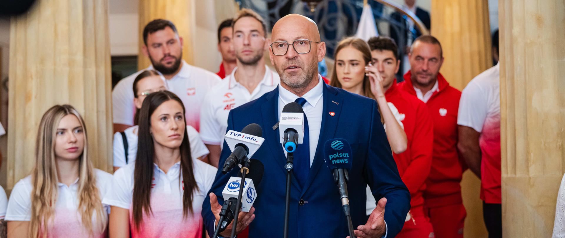Minister sportu i turystyki Jakub Rutnicki mówi do mikrofonów podczas konferencji prasowej. Za nim stoją reprezentanci Polski na mistrzostwa świata w lekkiej atletyce w Birmingham 2026.