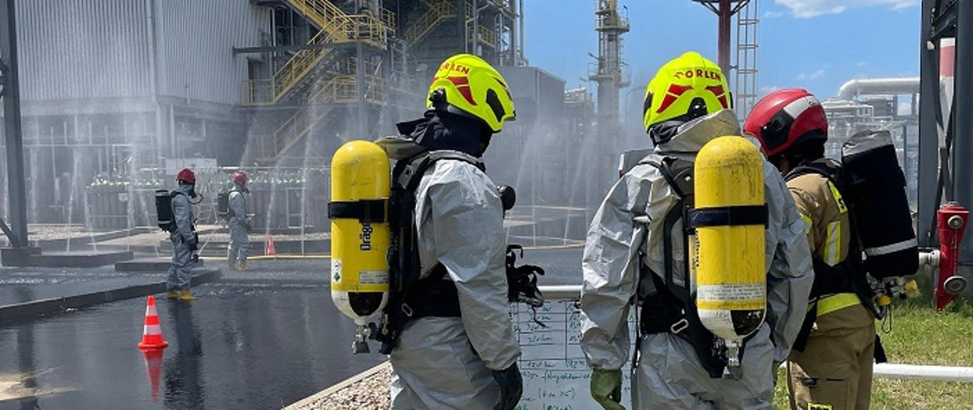 Na zdjęciu strażacy ubrani w specjalne kombinezony, posiadający na plecach aparaty dróg oddechowych realizujący założenie obrony instalacji.