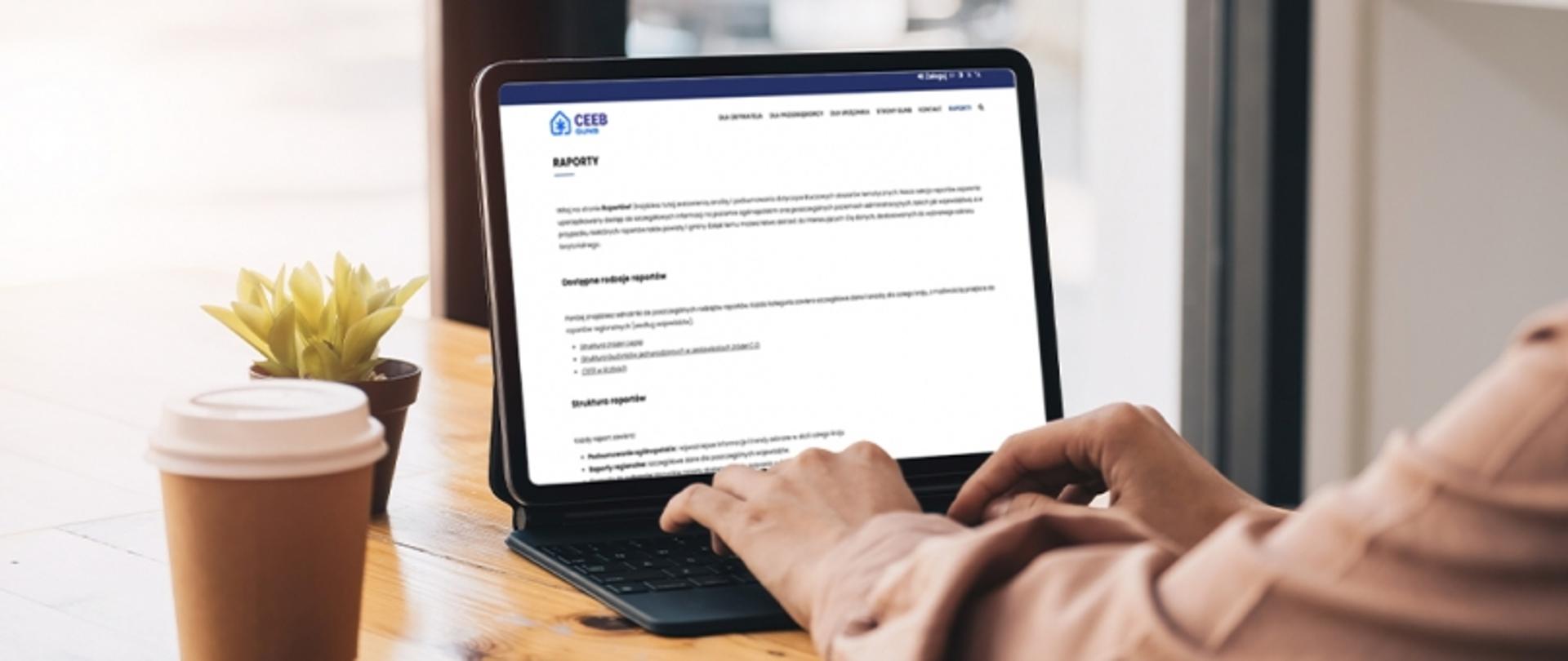 Osoba korzystająca z laptopa przy stoliku w kawiarni, przegląda zakładkę „Raporty” na stronie Centralnej Ewidencji Emisyjności Budynków (CEEB). Na stole stoją papierowy kubek z napojem i mała roślina w doniczce.
