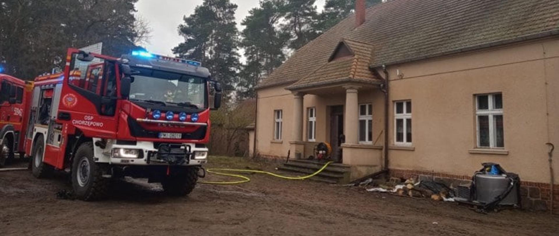 Zdjęcie przedstawia budynek mieszkalny i stojące obok samochody pożarnicze, które prowadzą działania podczas pożaru poddasza obiektu. W tle drzewa iglaste - sosny.