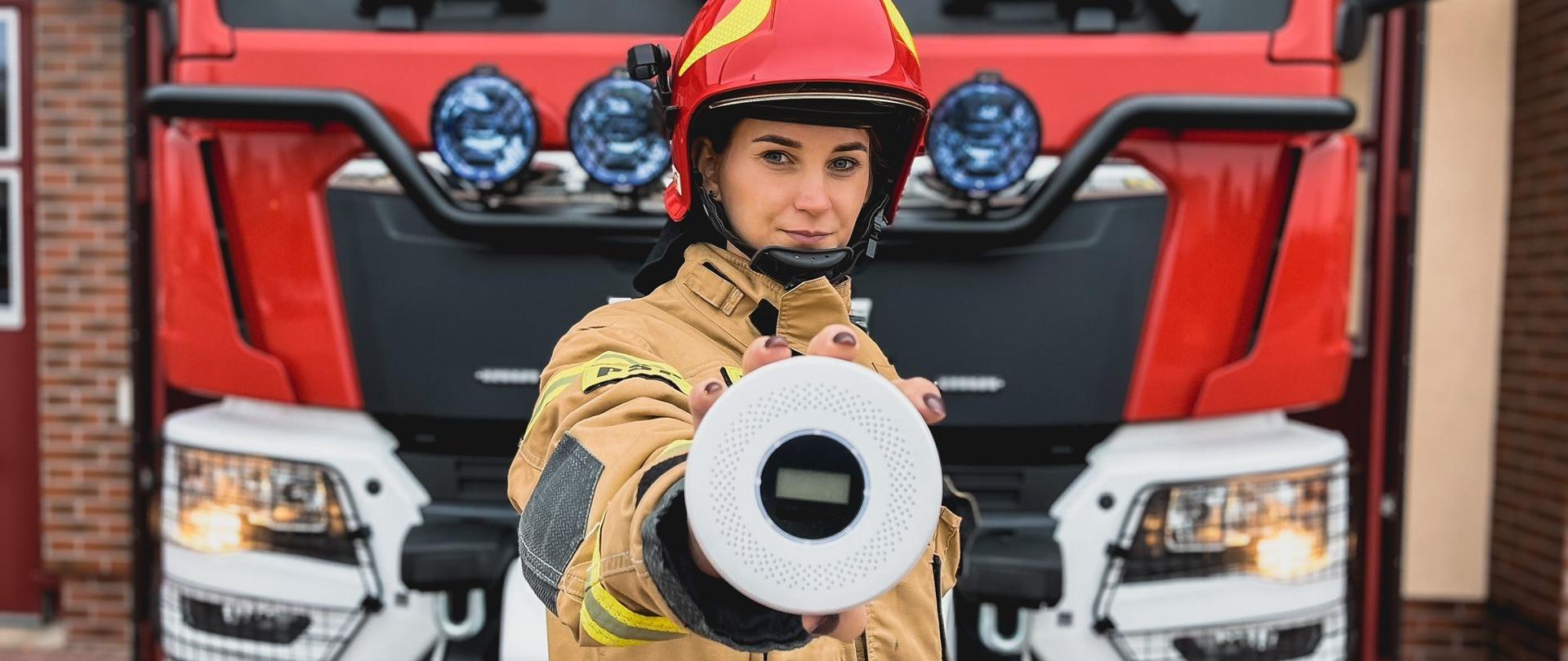 Strażak trzyma w ręku detektor tlenku węgla i dymu. W tle wóz strażacki i strażnica