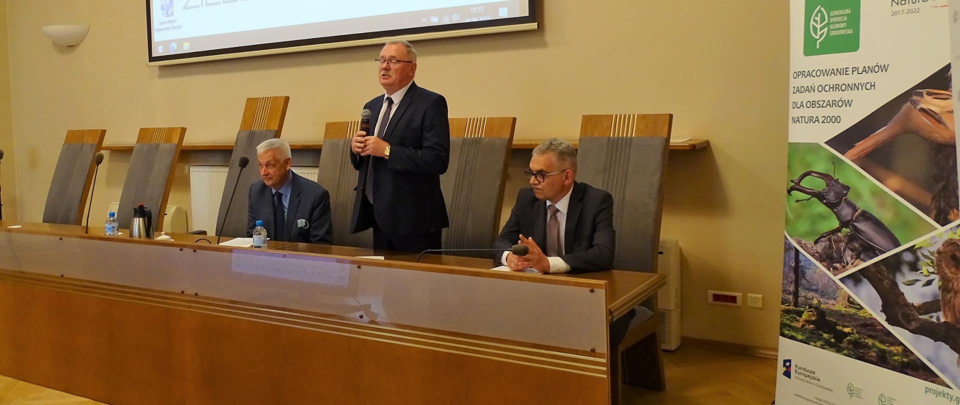 Przy stole trzech mężczyzn. Po lewej stronie siedzi mężczyzna ubrany w marynarkę koszulę i krawat po środku na stojąco przemawia Dyrektor RDOŚ ubrany w marynarkeęi krawat a po prawej siedzi Dyrektor Uniwersytetu Zielonogórskiego . Za nimi wyświetla się baner Uniwersytetu Zielonogórskiego. Po prawej stronie Baner RDOŚ