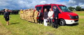 Zdjęcie 1.jpeg przedstawia oficjalną część wydarzenia. Na trawiastym placu, na tle czerwonego samochodu strażackiego marki Volkswagen, stoi grupa osób. Po lewej stronie w równym szeregu stoją strażacy w piaskowych (beżowych) mundurach koszarowych i czapkach z daszkiem. Przed nimi oraz obok pojazdu znajdują się zaproszeni goście oraz przedstawiciele władz i struktur strażackich, w tym kobieta w ciemnym garniturze, mężczyźni w strojach wizytowych oraz umundurowany oficer OSP w ciemnogranatowym mundurze wyjściowym z rogatywką. W tle widać zgromadzone drużyny oraz inne wozy bojowe.