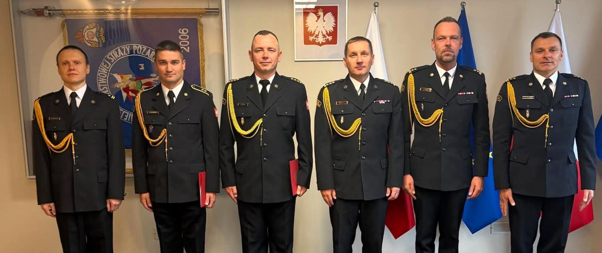 Sześciu strażaków w mundurach wyjściowych ze złotym sznurem podczas uroczystej odprawy w siedzibie komendy miejskiej.
