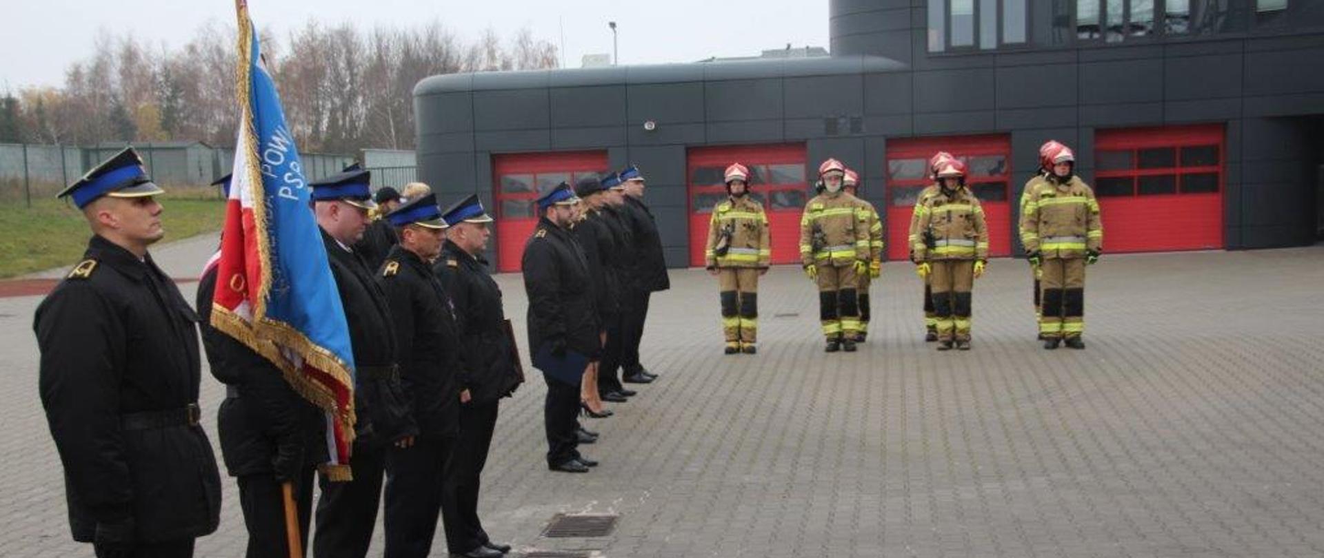 Na placu przed remizą stoją strażacy w szeregu. Na pierwszym planie poczet sztandarowy komendy, dalej załoga - w umundurowaniu wyjściowym, część w bojowym. W tle obiekt strażnicy.