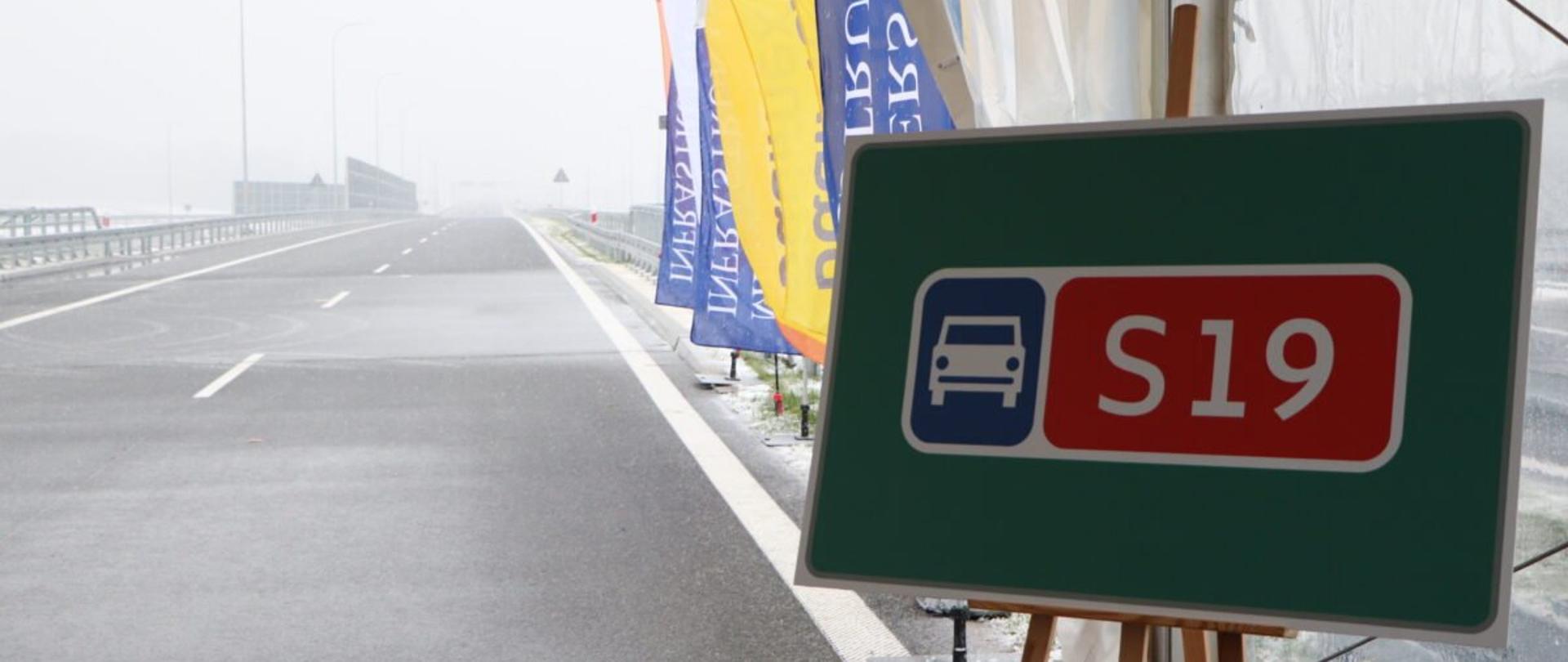 Zielona tablica na sztaludze, z napisem S19 i grafiką samochody na tle otwartego odcinka S19