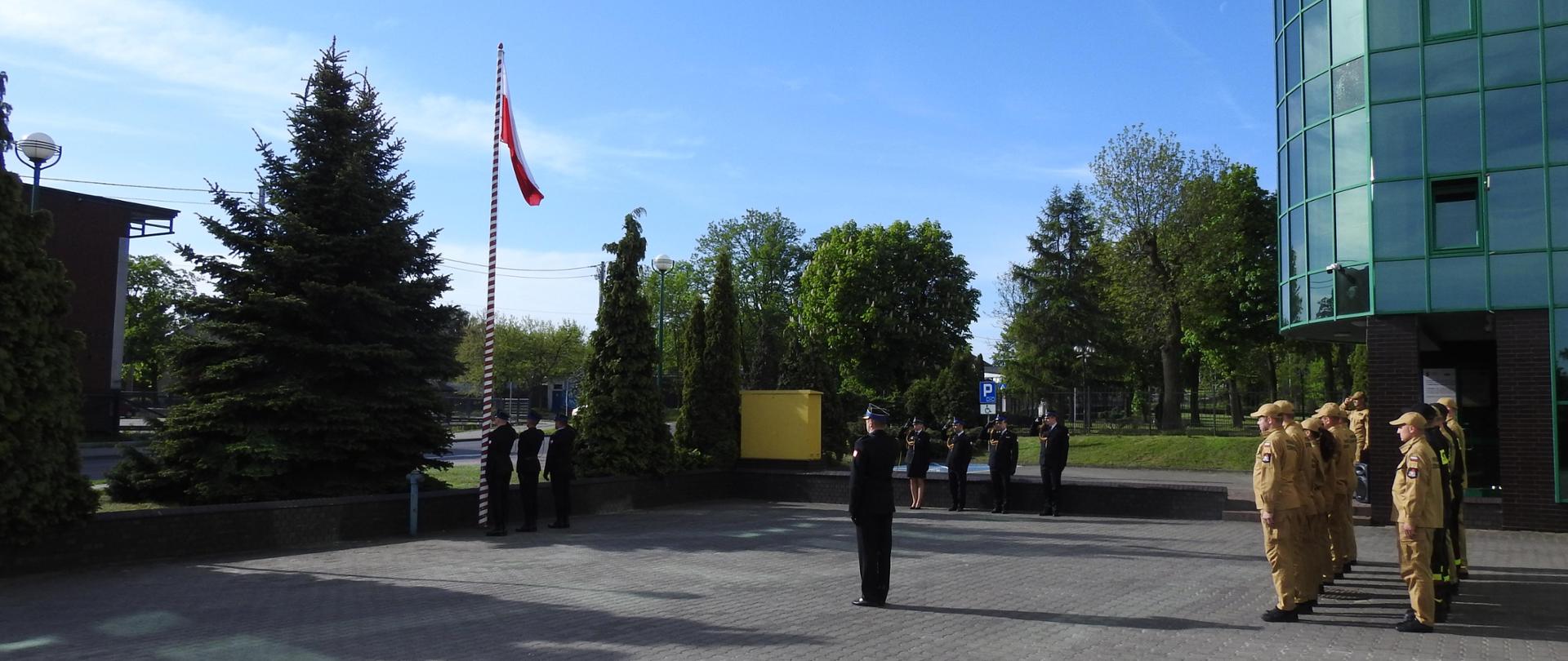 Strażacy stojący podczas uroczystej zbiórki w czasie podnoszenia flagi państwowej na maszt.