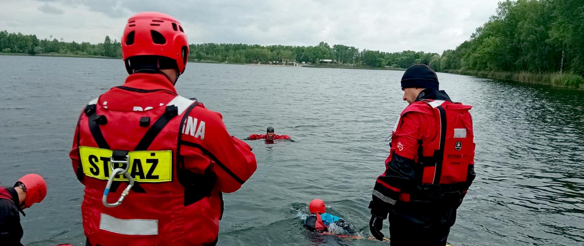 Na zdjęciu widać ćwiczenia strażaków z ratownictwa wodnego nad zbiornikiem wodnym Morawa w Katowicach. Kilka osób ubranych jest w czerwone kombinezony ratownicze i kaski ochronne. Jedna z osób na pierwszym planie ma na plecach napis „STRAŻ” – to strażak-ratownik. Stoi on na brzegu lub na pomoście, obserwując działania w wodzie. W wodzie znajdują się dwie osoby – jedna z nich płynie, ma na sobie suchy skafander. Druga osoba unosi się w wodzie nieco dalej, najprawdopodobniej też uczestnicząc w ćwiczeniach ubrana także w suchy skafander. W tle widoczne jest spokojne zbiornik wodny, otoczone zielonym lasem. Niebo jest zachmurzone.
