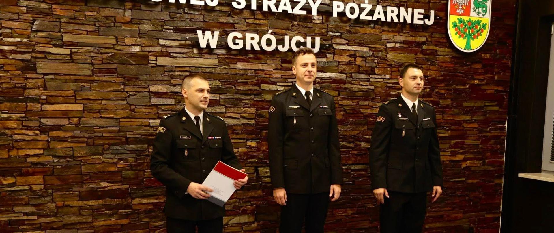 Na pierwszym zdjęciu widzimy formalną uroczystość, najprawdopodobniej związaną z KP Państwowej Straży Pożarnej w Grójcu. Na pierwszym planie znajdują się trzej mężczyźni w mundurach galowych. Stoją naprzeciwko siebie w poważnej postawie — to moment mianowania dowódcy JRG