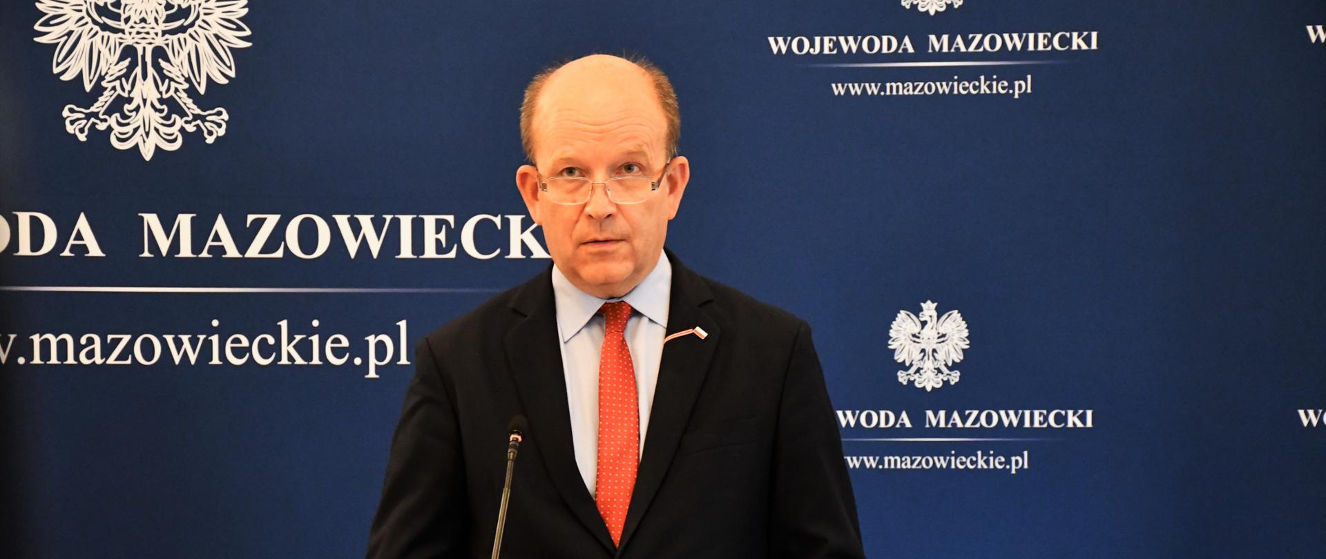 Zdjęcie przedstawia elegancko ubranego mężczyznę - w ciemną marynarkę, błękitną koszulę i czerwony krawat. Mężczyzna ten stoi za mównicą, na tle ścianki granatowej ścianki wizerunkowej. Na ściance tej znajduje się logo Wojewody (zawierające Godło Polski oraz napis "Wojewoda Mazowiecki")