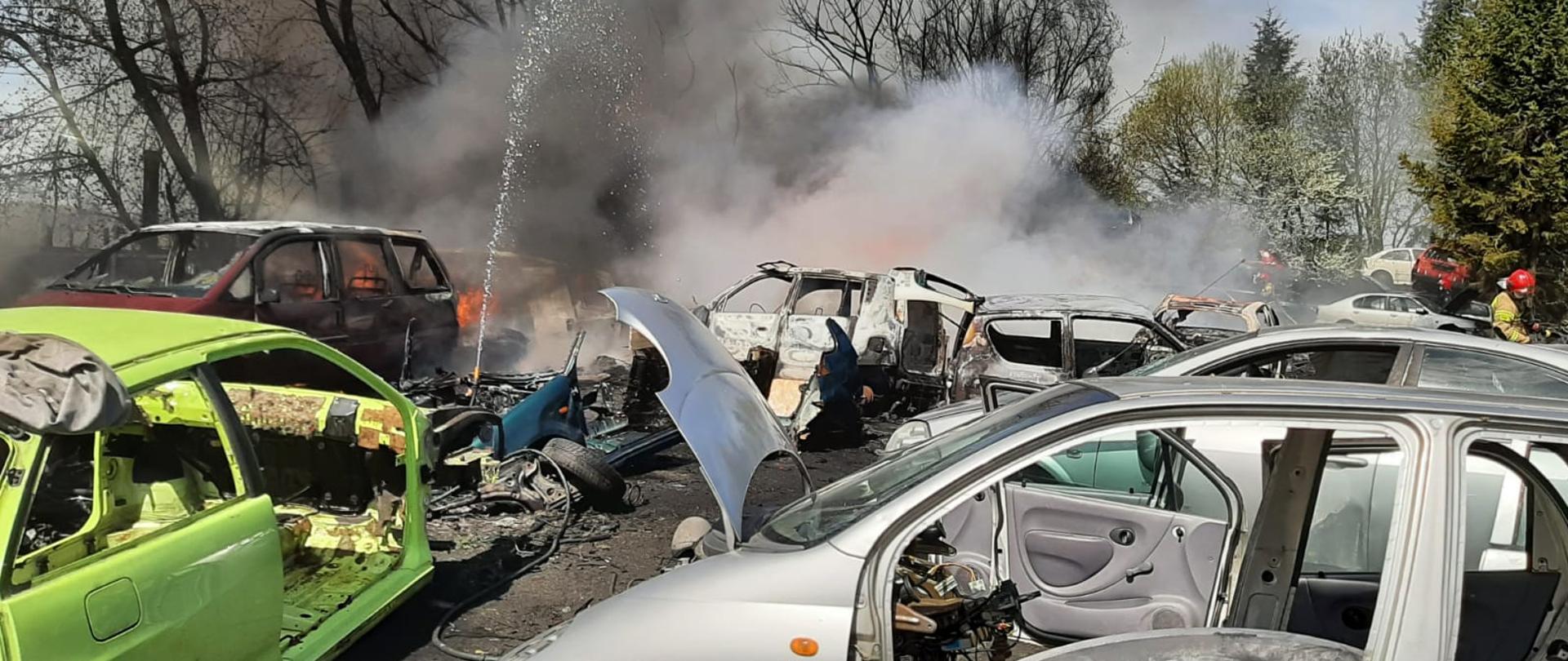 Złomowisko pojazdów samochodowych, na pierwszym planie nadpalone elementy karoserii samochodów osobowych, w tle kłęby dymu. 