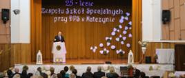 Minister Czarnek stoi przy mównicy i mówi do ludzi siedzących na ustawionych w rzędy krzesłach. Za nim złota kotara a na niej niebieski napis 25-lecie Zespołu Szkól specjalnych przy DPS w Matczynie.