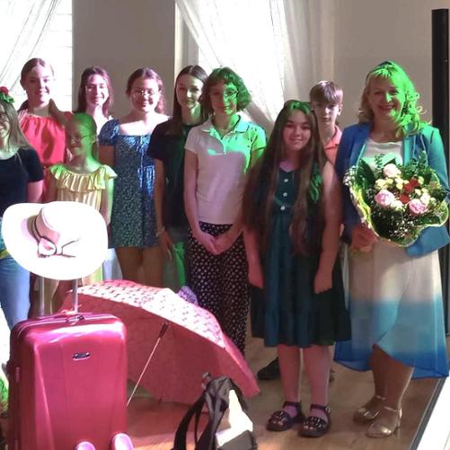 Zdjęcie zbiorowe na scenie przy fortepianie. Dziewczęta i chłopiec kolorowo ubrani stoją ustawieni w dwóch rzędach. Przed nimi walizki, klapki plażowe, piłka, hulajnoga. Nauczycielka trzyma w ręku kwiaty.