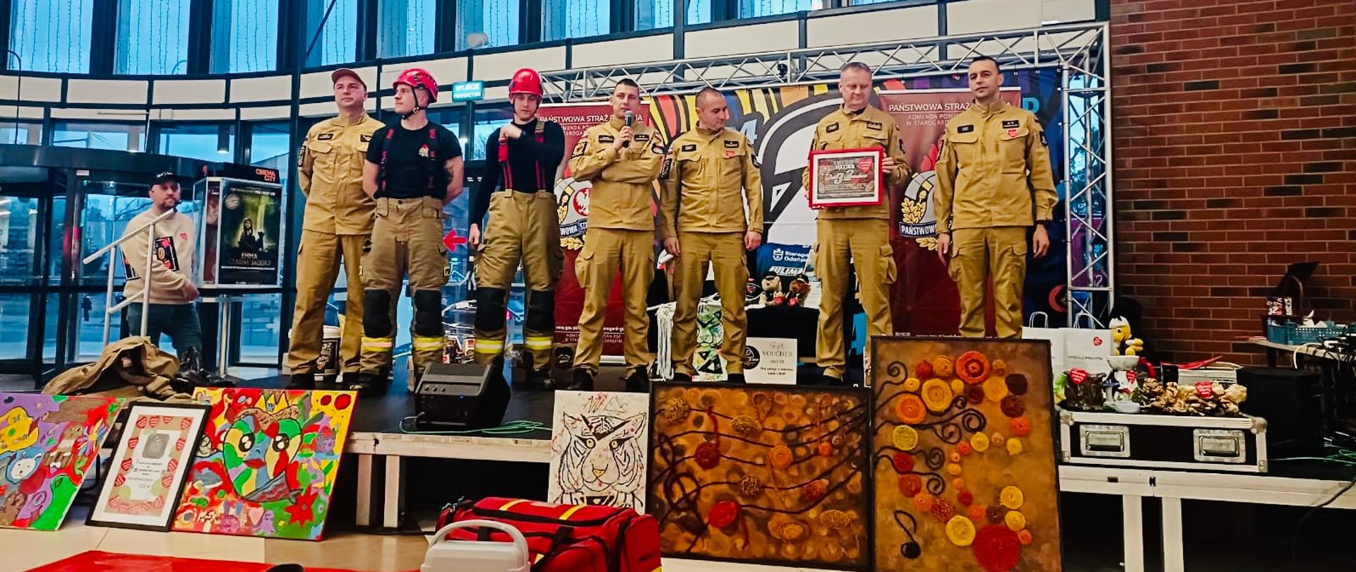 Zdjęcie strażaków i kadry kierowniczej Komendy uczestniczących w pokazie i licytacji WOŚP, Komendant trzyma licytowany voucher