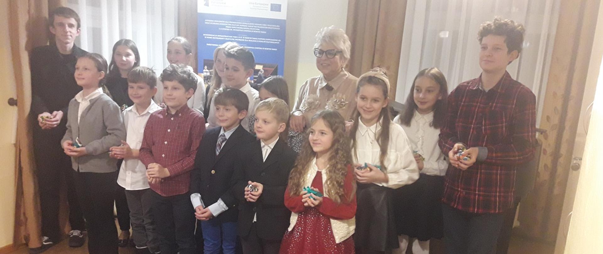 Zdjęcie grupowe. Dzieci biorące udział w audycji wraz z nauczycielką stoją w dwóch rzędach i trzymają w swych dłoniach bombki choinkowe. Na pierwszym planie puste krzesła a na jednym z nich słodycze. Za nimi w tle dwa okna przysłonięte firankami i zasłonami oraz baner unijny. Pod sufitem zwisające dwie zapalone lampy.