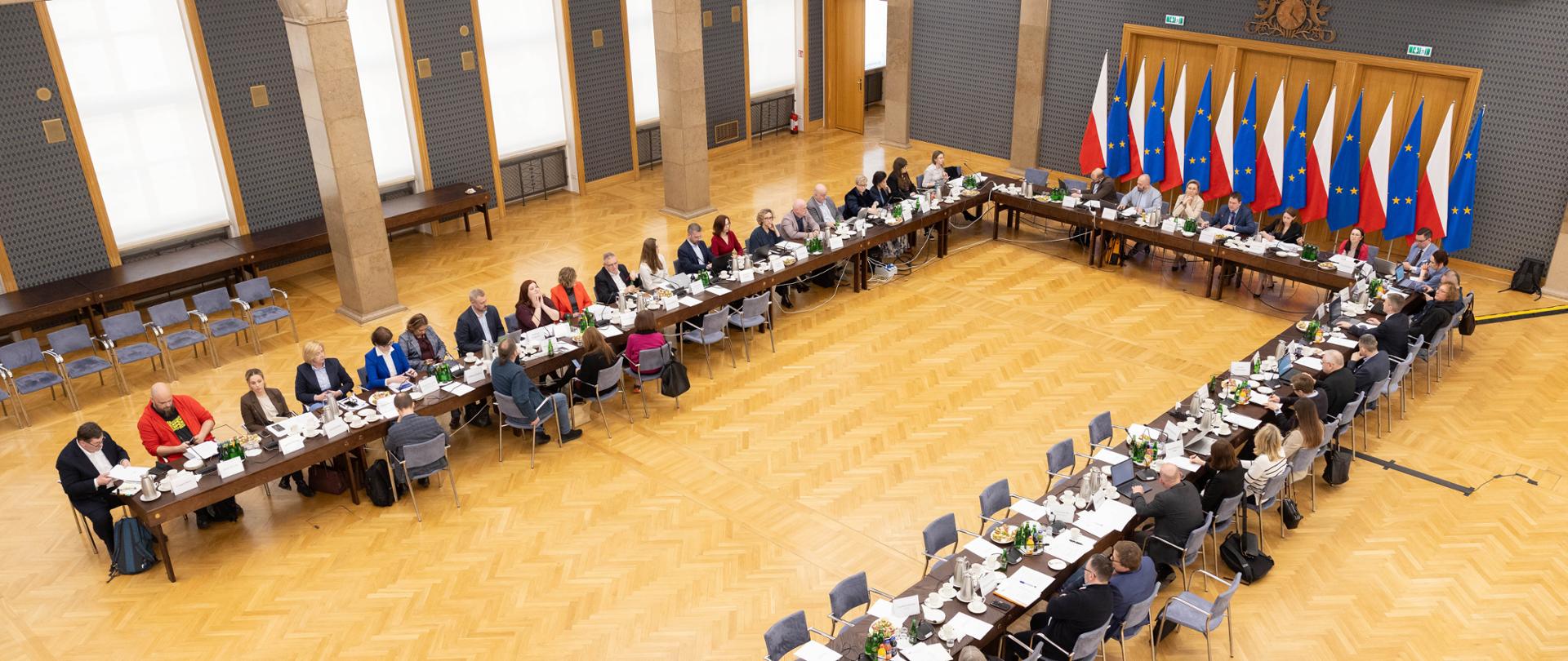 Szeroki kadr obejmuje salę konferencyjną. Na zdjęciu uczestnicy posiedzenia obradują przy stołach konferencyjnych ustawionych w literę U. U szczytu stołu między innymi Pani Adriana Porowska, Sekretarz Stanu w KPRM, Wiceprzewodnicząca Komitetu ds. Pożytku Publicznego. W tle flagi Polski i Unii Europejskiej.