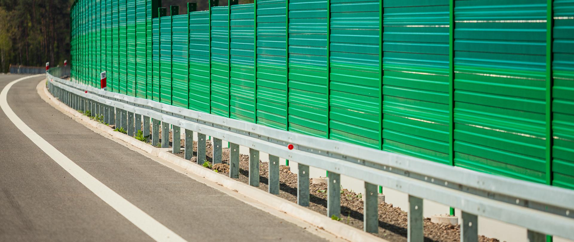 Na zdjęciu widoczna jest asfaltowa droga biegnąca równolegle do wysokiego, zielonego ekranu akustycznego. Ekran składa się z metalowych paneli zamocowanych na pionowych słupkach, a od jezdni oddziela go metalowa barierka ochronna. W tle rozciąga się gęsty las, a nad nim rozpościera się jasne, bezchmurne niebo, co sugeruje pogodny dzień.