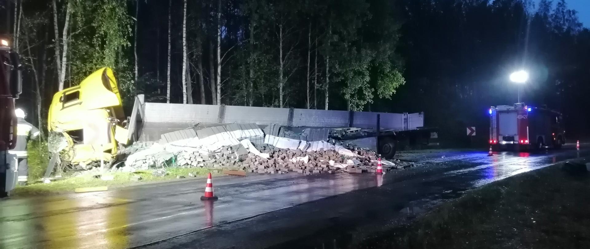 Zdjęcie przedstawia ciężarówkę, która leży w rowie,
z przyczepy wysypana jest kostka brukowa która zajmuje plowę jezdni.
Zdjęcie wykonane jest w porze wieczorowej.
Ciężarówka z dwóch stron oświtlona jest reflektorami.
Widać, że niedawno padał deszcz ponieważ jezdnia lśni się połyskującą taflą.
Droga jest całkowicie nieprzejezdna wzdłuż samochodu leżącego w rowie ustawione
są dwa pachołki ostrzegawcze uniemożliwijące poruszanie się.