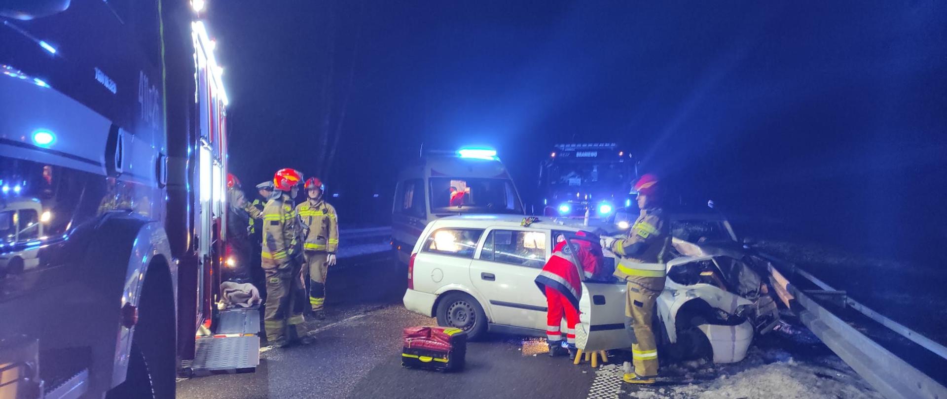 Zdjęcie zrobione nocą. W poprzek drogi z prawej strony białe auto kombi rozbity przód o barierki. Z lewej strony i w oddali samochody strażackie i Karetka. Samochód strażacki oświetla teren. Przy samochodzie strażacy w czerwonych hełmach i ratownik w czerwonej kurtce i spodniach zagląda do uszkodzonego białego pojazdu.