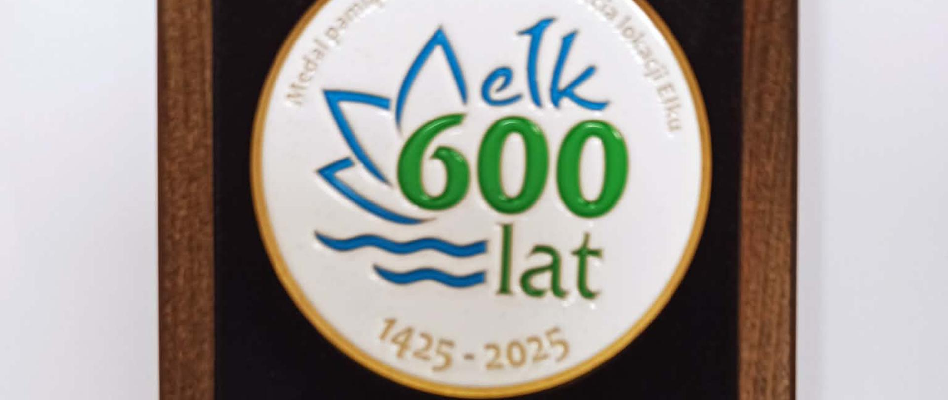 Zdjęcie przedstawia pamiątkowy medal umieszczony w drewnianej ramce oraz certyfikat. Na medalu widnieje napis: ‘Ełk 600 lat’ oraz daty ‘1425–2025’, a także symbol graficzny miasta. Poniżej znajduje się certyfikat. Treść certyfikatu brzmi: ‘Z dumą przekazuję medal pamiątkowy, który został wybity z okazji 600-lecia lokacji Ełku (1425–2025). Niech będzie świadectwem szacunku dla przeszłości oraz źródłem dumy i inspiracji na kolejne stulecia.’ Dokument podpisany przez Tomasza Andrukiewicza, Prezydenta Miasta Ełku. Na dole certyfikatu umieszczone jest logo ‘Ełk 600 lat’.”