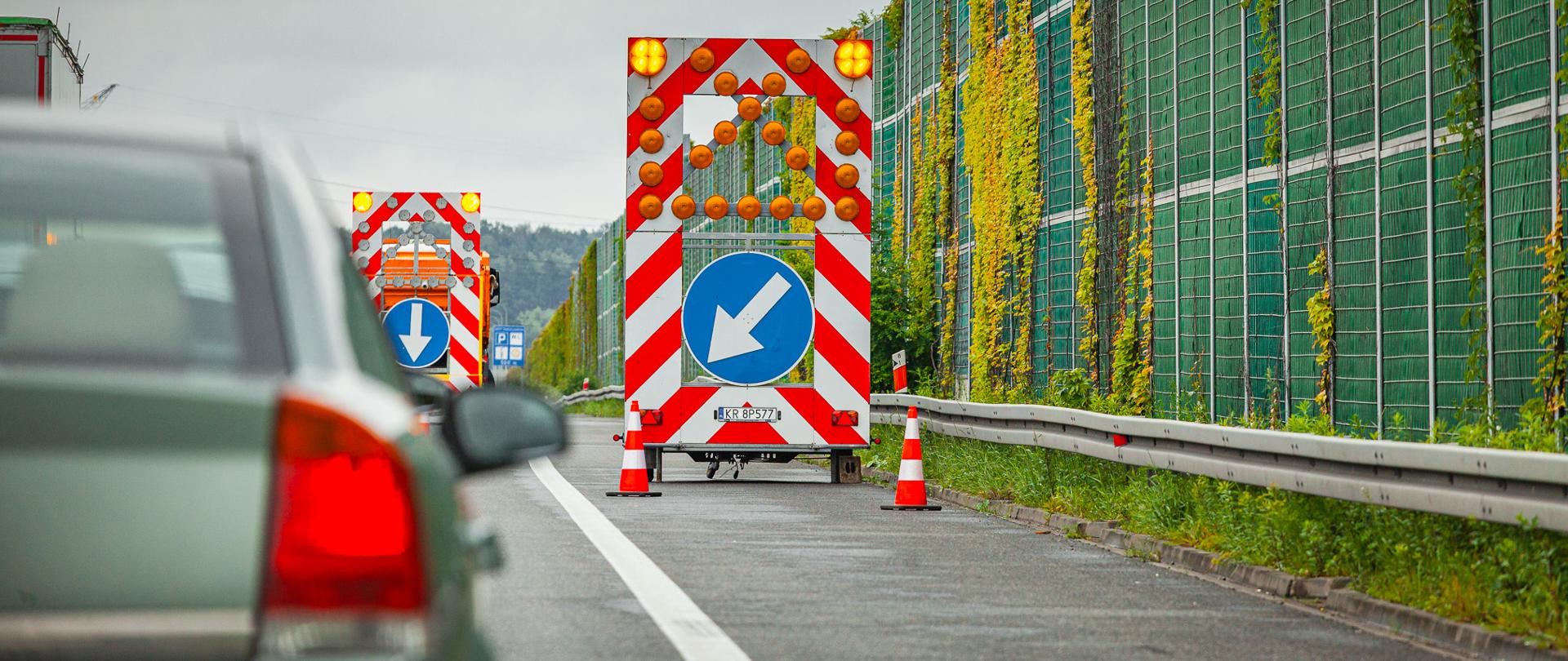 Prace drogowe na autostradzie A1. Na pasie awaryjnym ustawiono pojazdy z tablicami świetlnymi ostrzegającymi kierowców o zwężeniu jezdni i konieczności zmiany toru jazdy. Obok przebiegają ekrany akustyczne porośnięte pnączami, a w kadrze widoczny jest również przejeżdżający samochód.