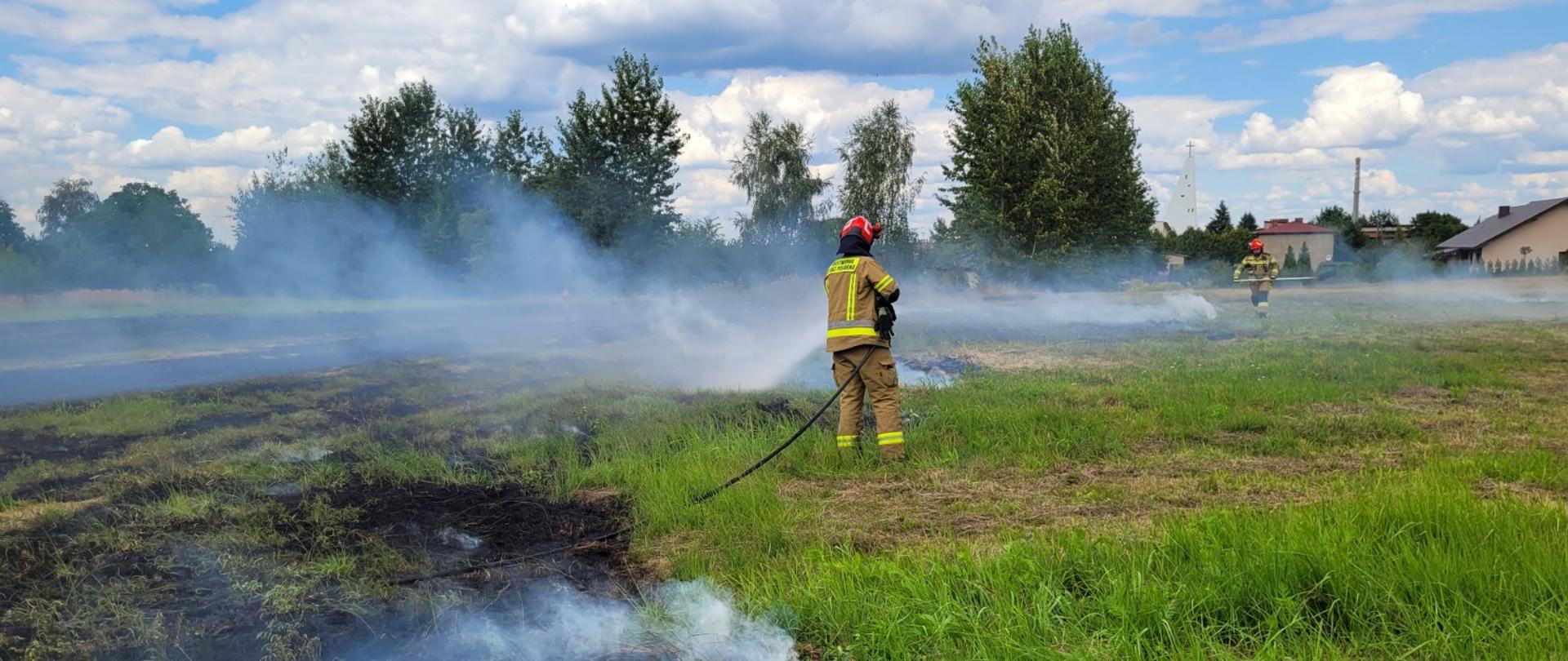 Zdjęcie przedstawia płonącą łąkę z pozostałościami siana. Dwóch strażaków gasi. W tle wieża kościoła i domy mieszkalne i drzewa.