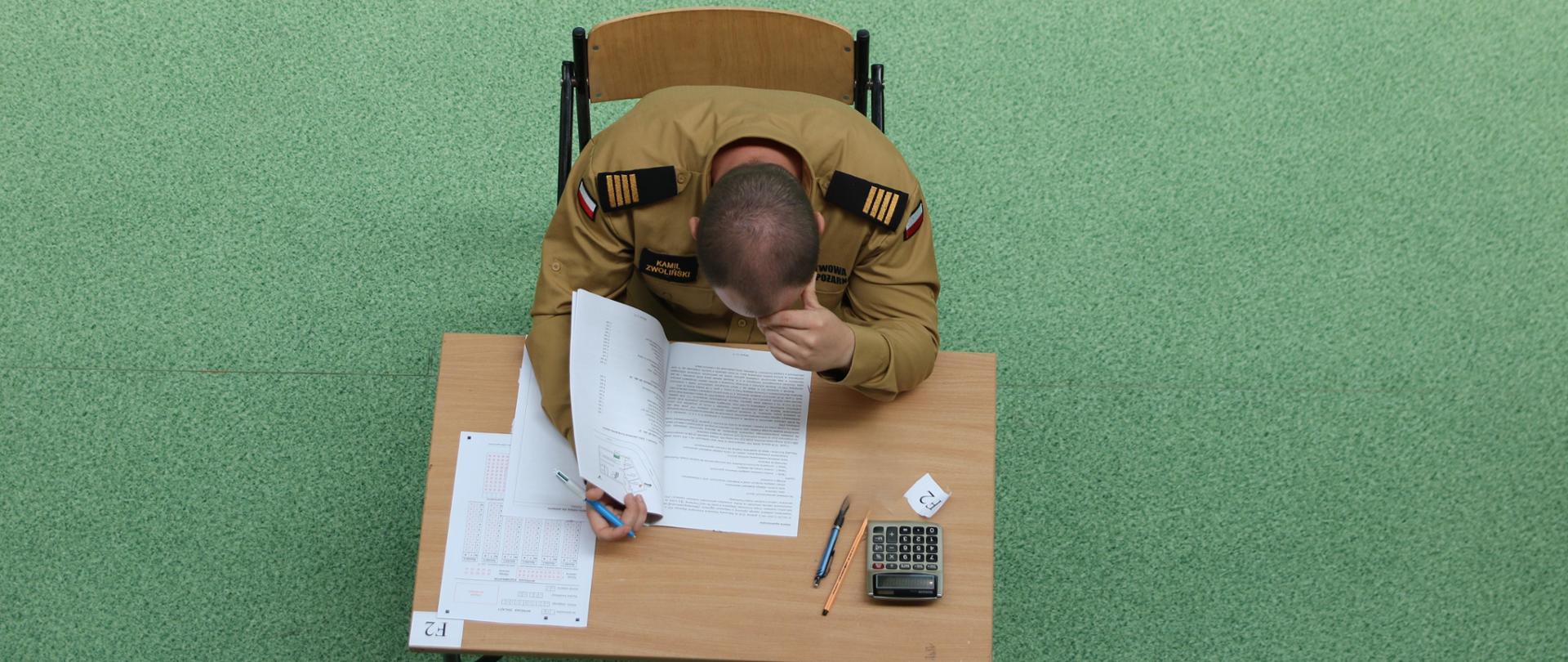Siedzący przy stoliku mężczyzna. Strażak w umundurowaniu służbowym. Pochyla się nad kartką z zadaniem egzaminacyjnym. 