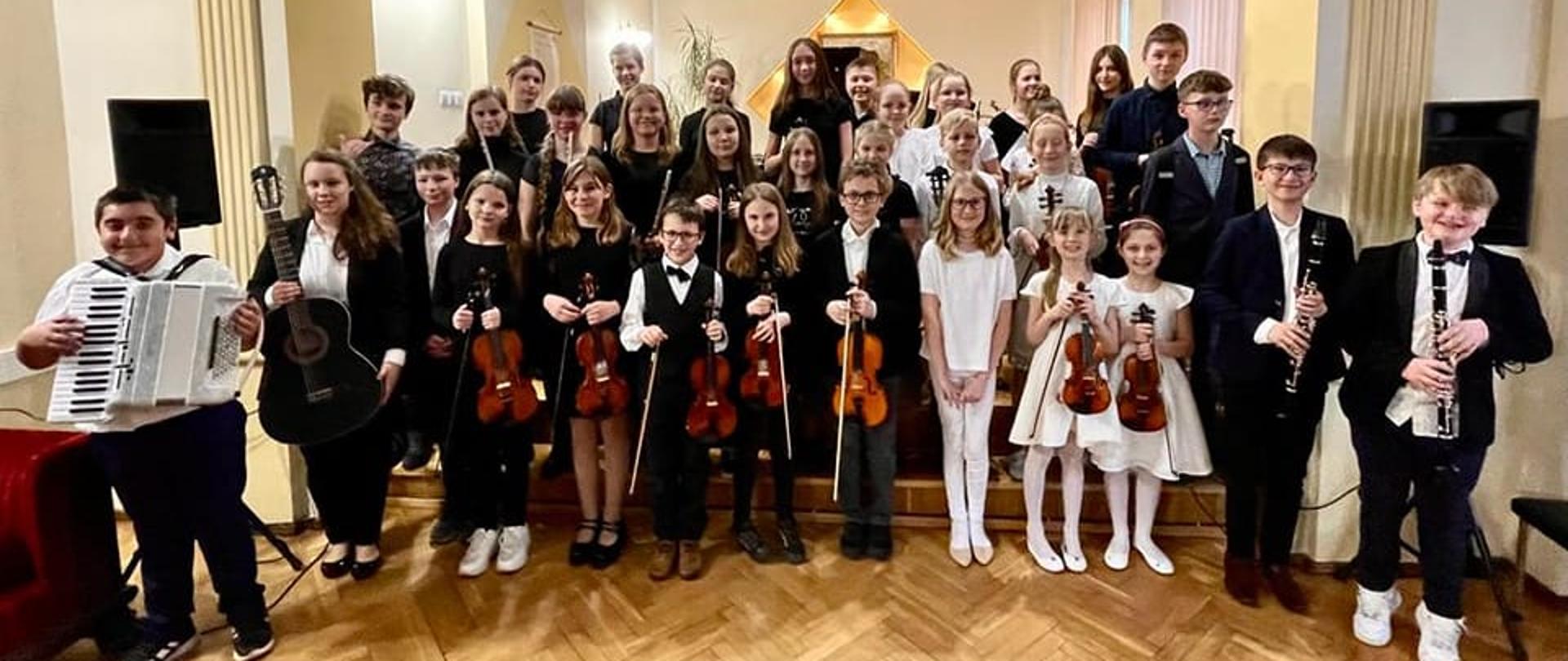 Na zdjęciu grupowym wszyscy uczestnicy koncertu zespołów kameralnych ubrani w eleganckie stroje i trzymający swój instrument
