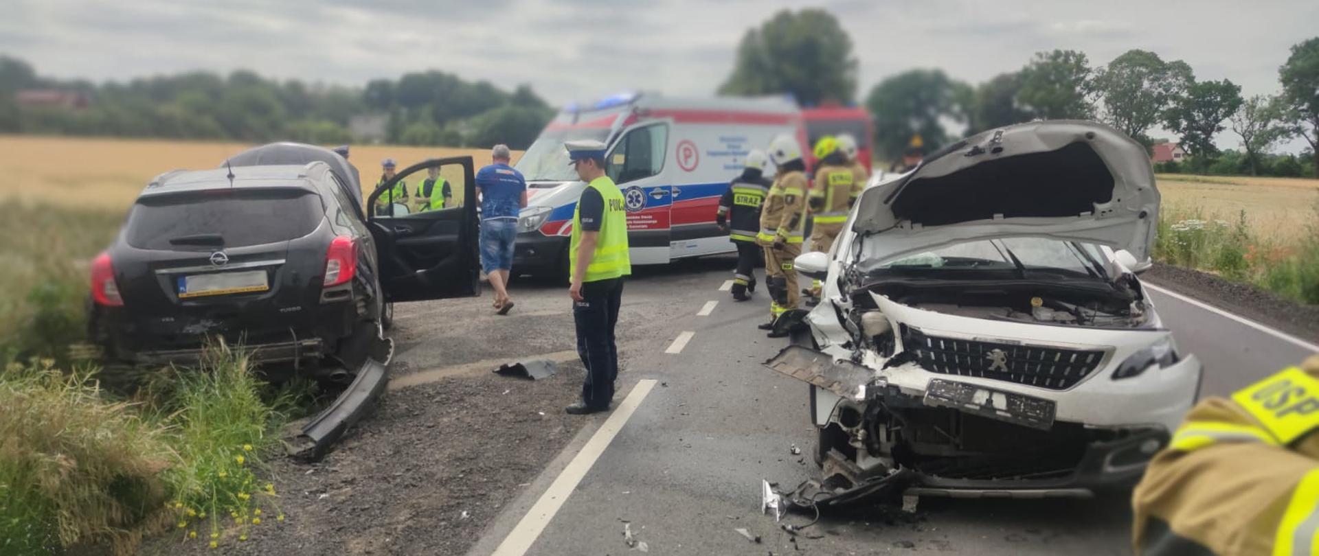 na drodze stoi rozbity biały samochód, z lewej na poboczu czarny, pomiędzy samochodami stoi policjant dalej grupa strażaków, karetka pogotowia oraz samochód pożarniczy.