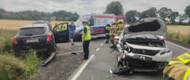 na drodze stoi rozbity biały samochód, z lewej na poboczu czarny, pomiędzy samochodami stoi policjant dalej grupa strażaków, karetka pogotowia oraz samochód pożarniczy.