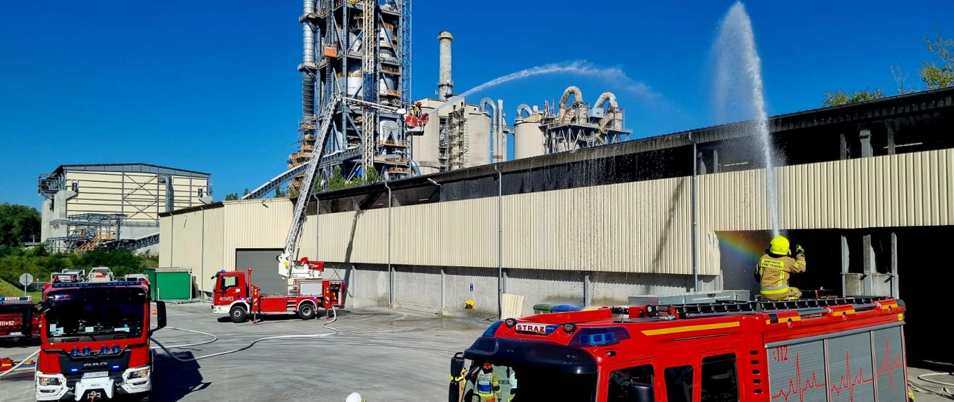 Zdjęcie obrazuje dwa samochody gaśnicze oraz samochód operacyjny PSP. Strażacy prowadzą działania gaśnicze podczas prowadzonych ćwiczeń. Podają dwa prądy wody: jeden z podnośnika hydraulicznego z wysokości, drugi z działka wodno-pianowego.